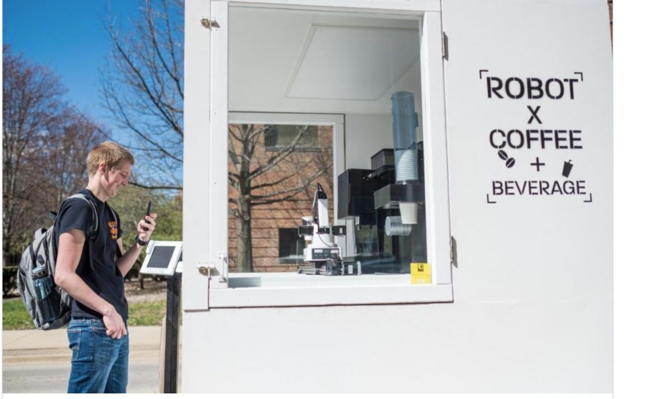 A student buys coffee at the trailer window while the robot arm works inside