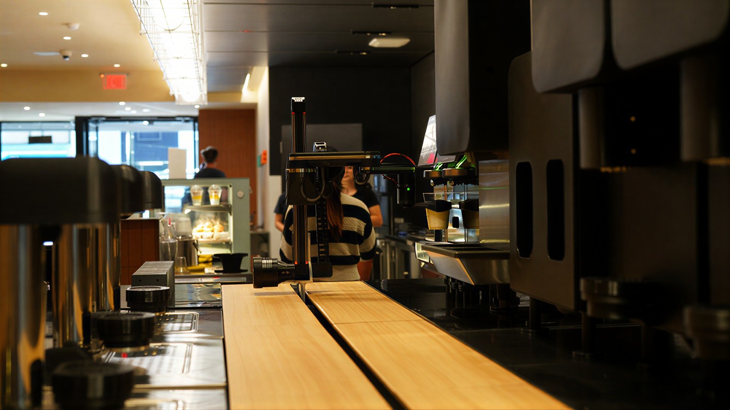 Gantry arm at the bar: machine column, camera head, cups filling over the wood counter