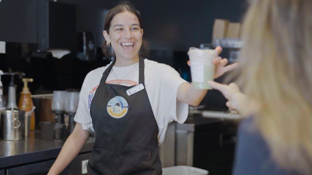 A barista in a Yummy Future apron laughing as she hands a cup across the counter to a customer