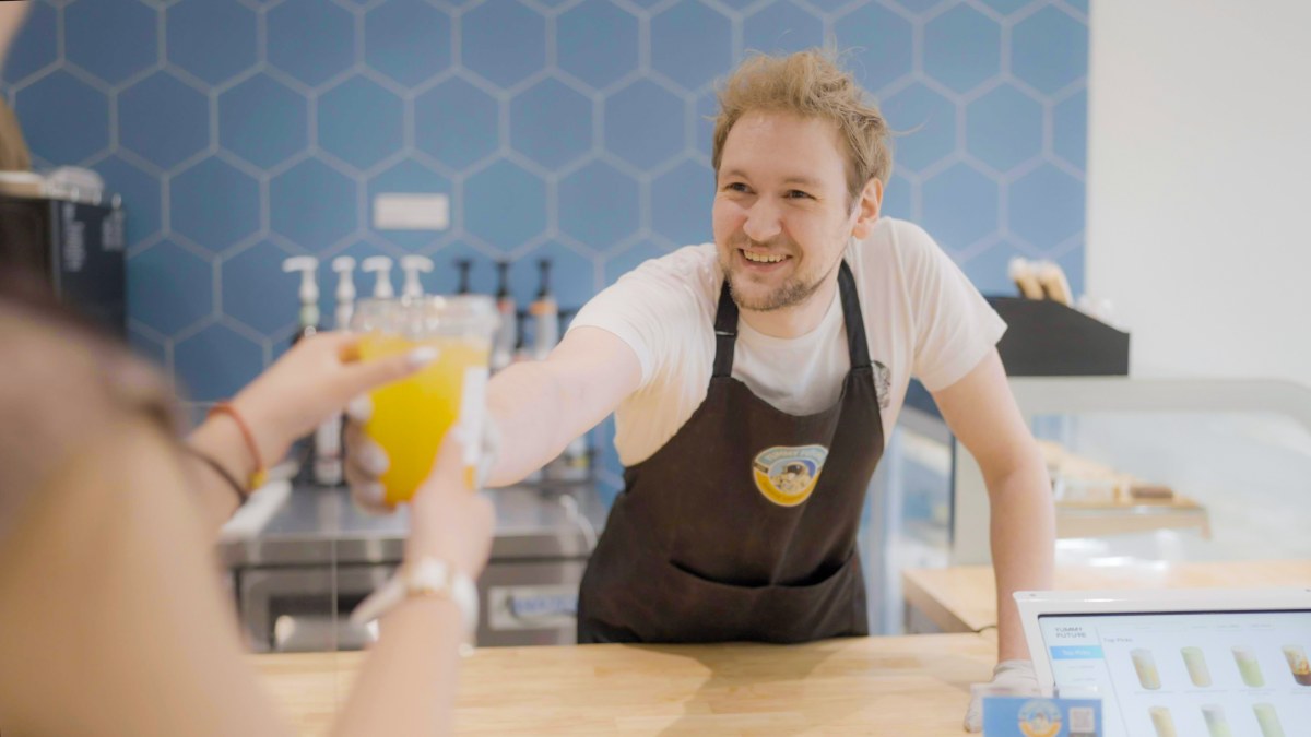 Barista laughs while handing a cup of fresh orange juice across the counter to a customer